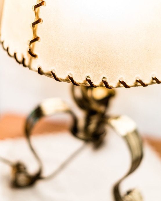 Nahaufnahme einer rustikalen Tischlampe mit genähtem Lederschirm und geschwungenem Metallfuß.