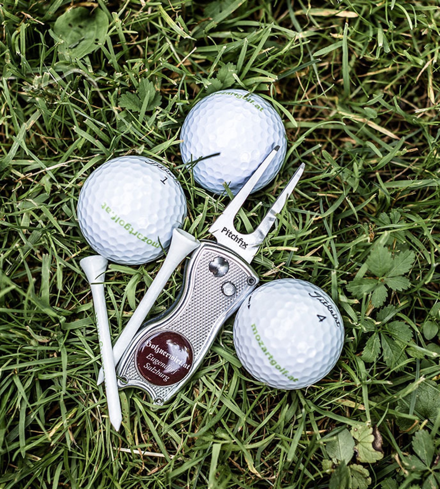 Drei Golfbälle, zwei Tees und eine Pitchgabel der Marke Pitchfix auf grünem Gras von oben fotografiert.