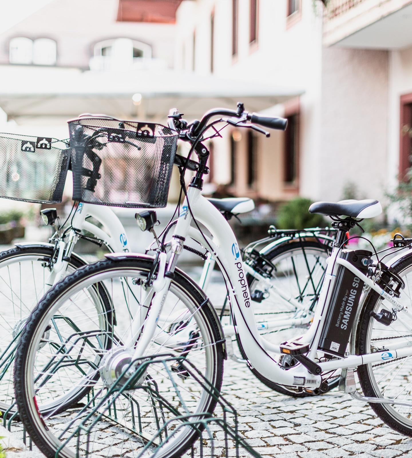 Zwei weiße Prophete E-Bikes mit Samsung SDI Akku parken in einem Fahrradständer auf Kopfsteinpflaster.