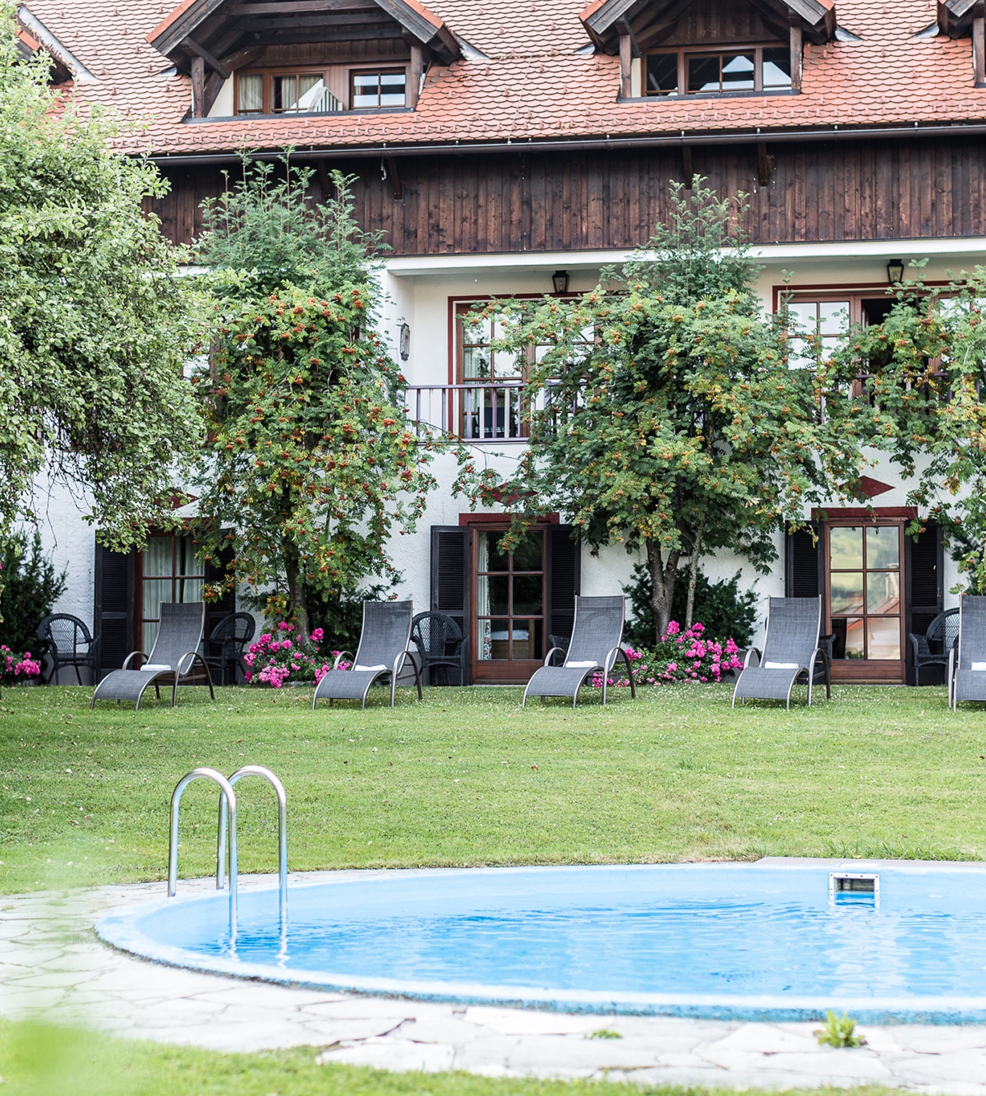 Außenpool mit Liegestühlen auf einer Wiese vor einem rustikalen Hotel mit Bäumen und Blumen.
