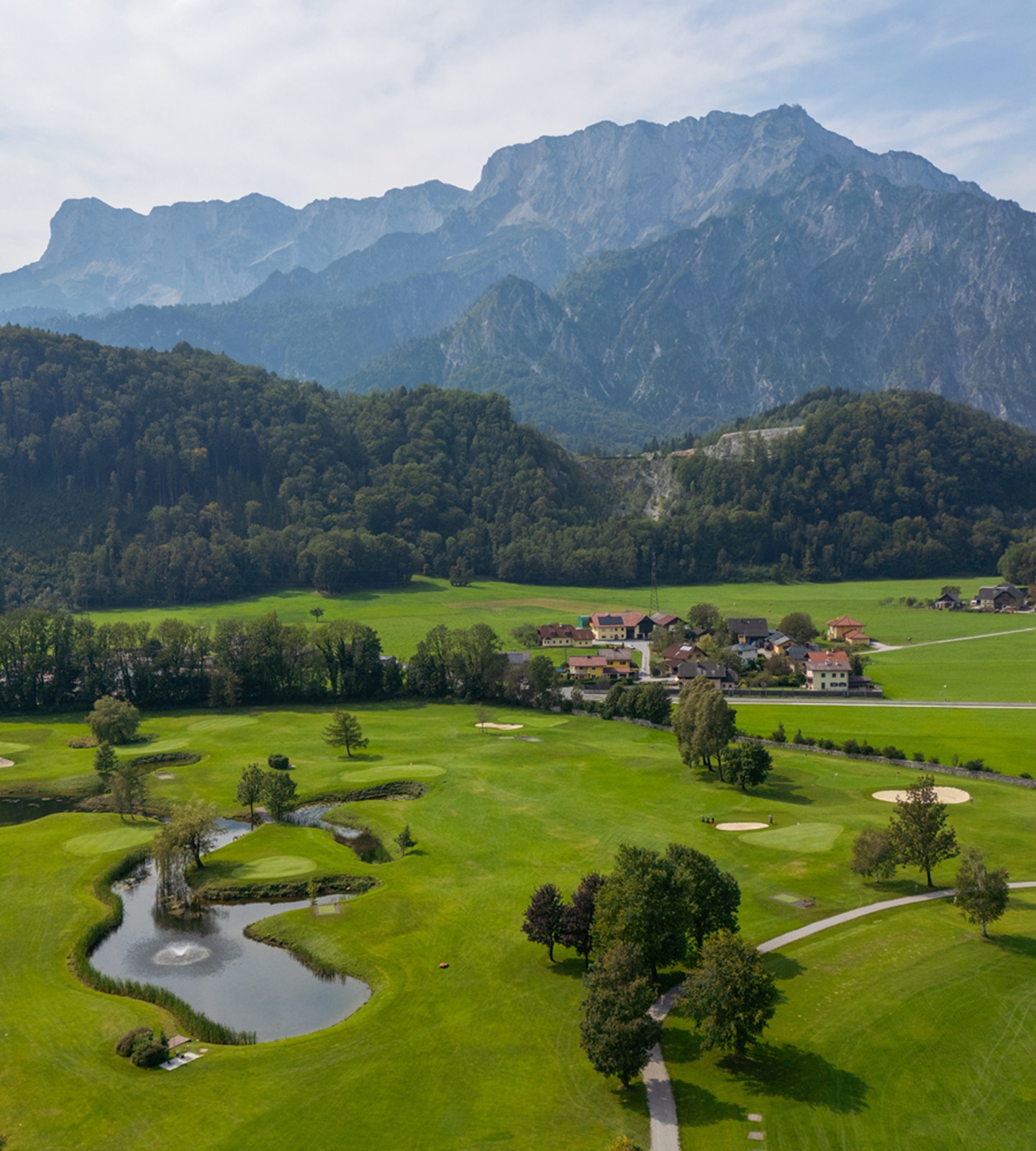 Luftaufnahme eines grünen Golfplatzes mit Teich vor einer bewaldeten Hügellandschaft und hohen Bergen.