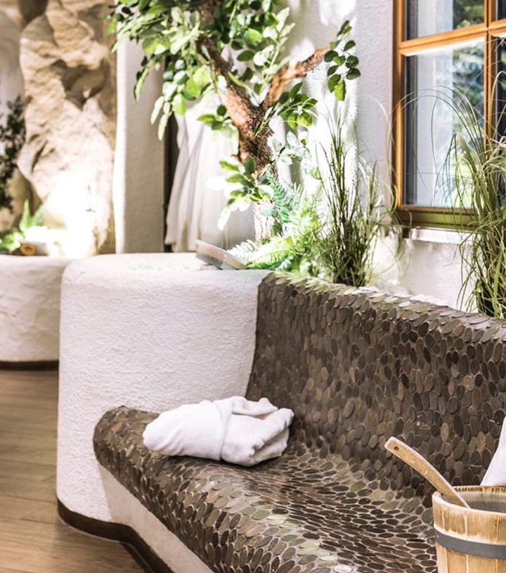 Mosaik-Sitzbank mit Bademantel und Saunaeimer in einem hellen Wellnessbereich mit Pflanzen.