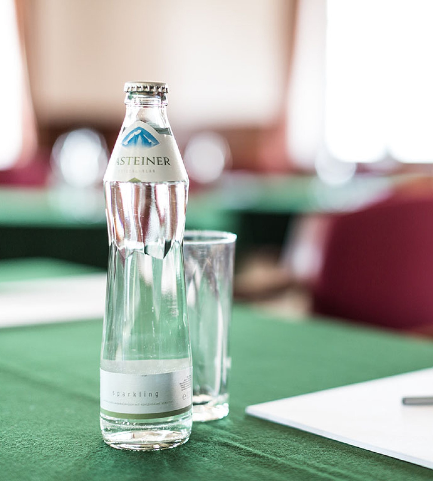 Eine Flasche Gasteiner Sparkling Mineralwasser und ein leeres Glas stehen auf einem grünen Tisch in einem Seminarraum.
