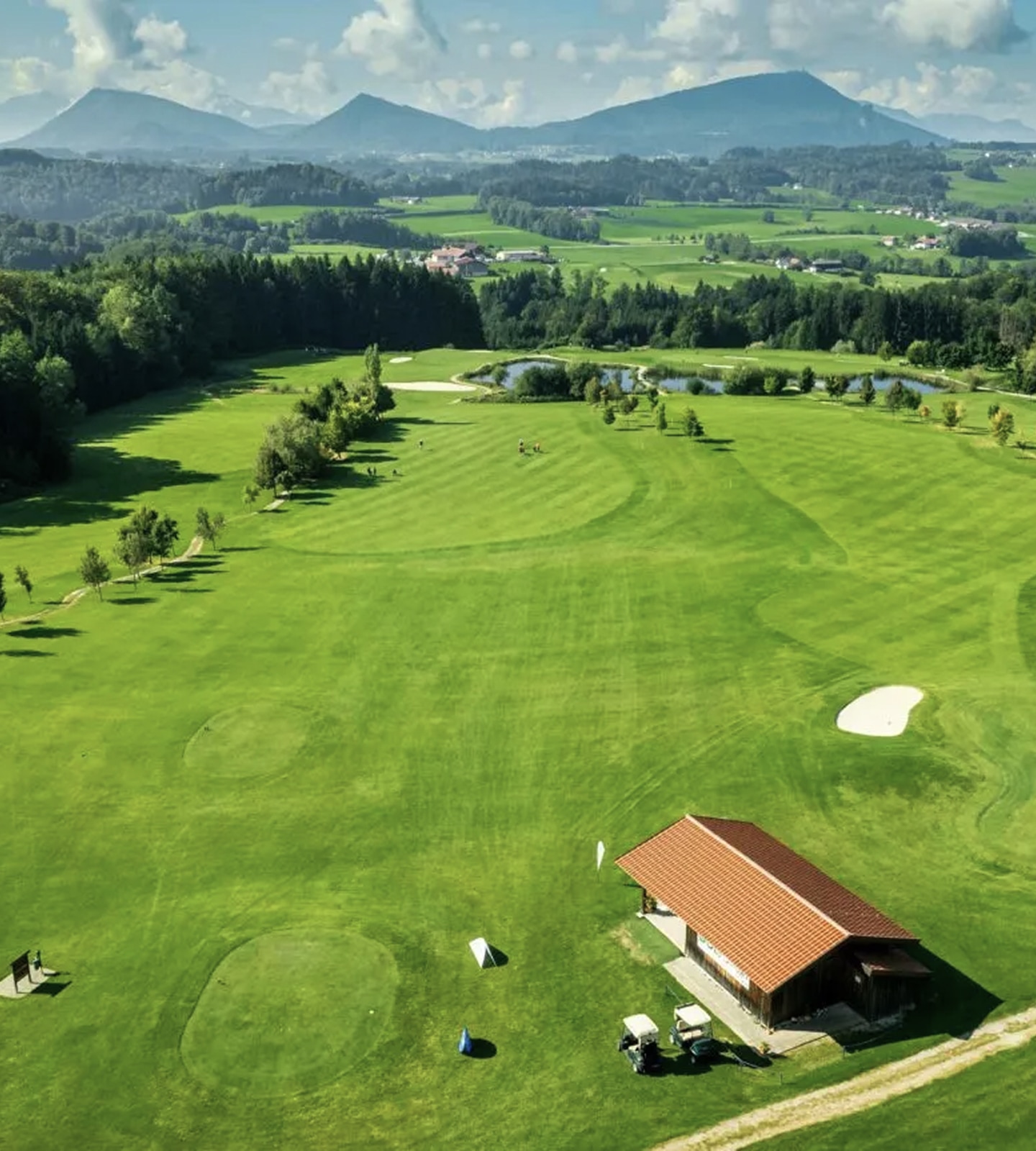 Luftaufnahme eines grünen Golfplatzes mit Hütte, Carts und Bergen im Hintergrund.