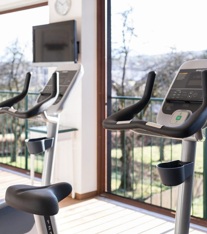 Zwei Ergometer stehen in einem hellen Fitnessraum vor bodentiefen Fenstern mit Blick ins Grüne.