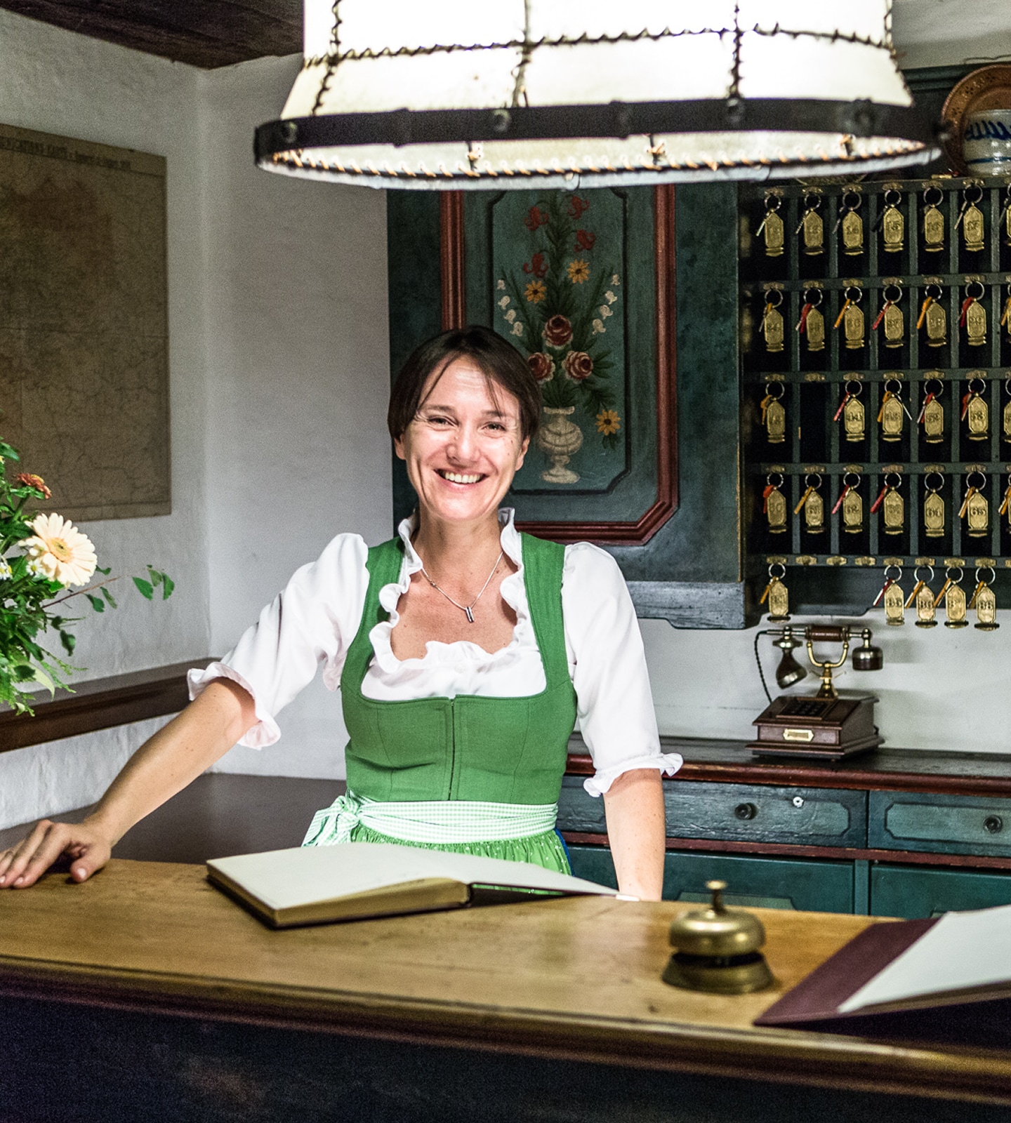 Eine lächelnde Frau im Dirndl steht an einer rustikalen Hotelrezeption mit Schlüsselbrett im Hintergrund.