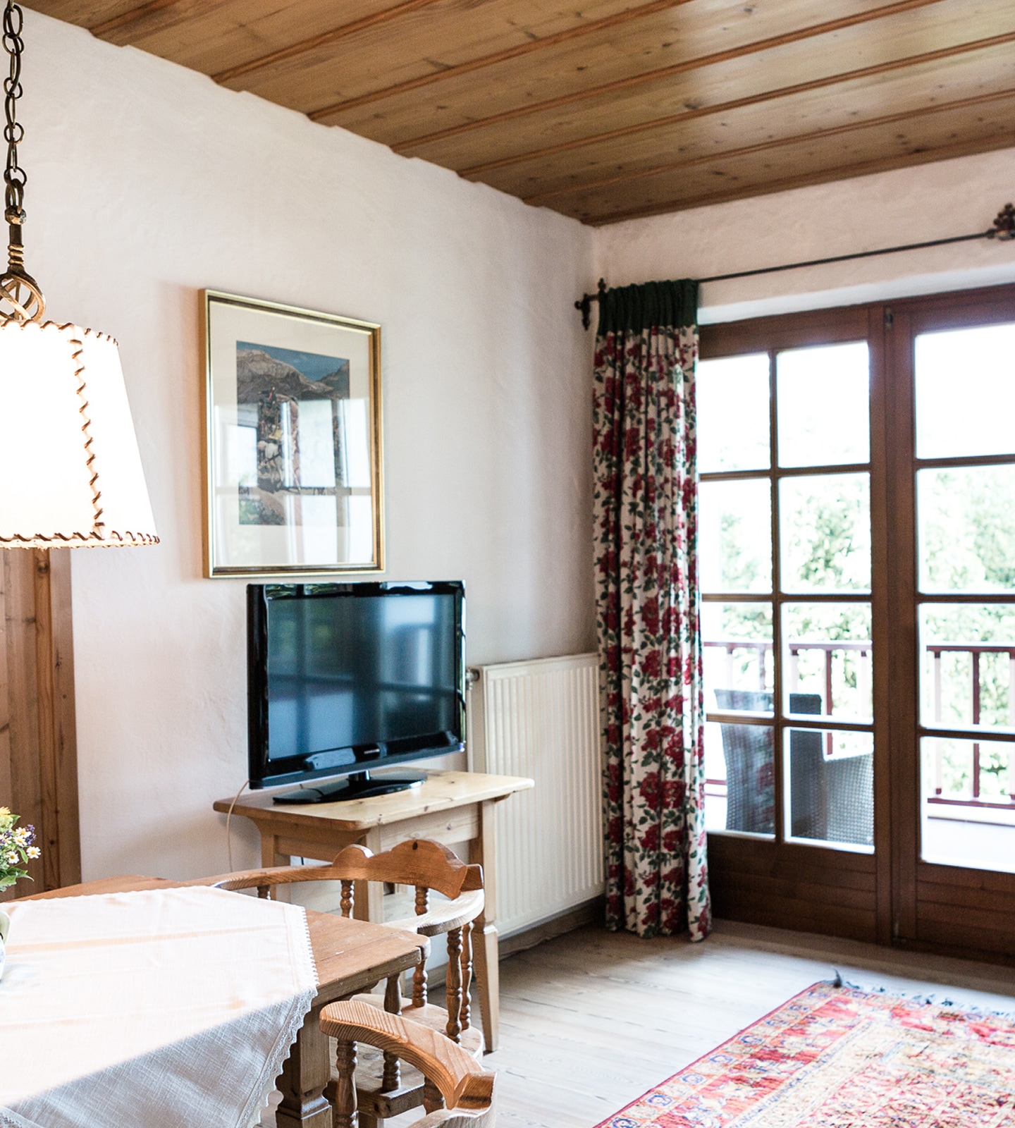 Gemütliches Wohnzimmer mit Holzdecke, Fernseher auf Holztisch und einer Balkontür mit geblümten Vorhängen.