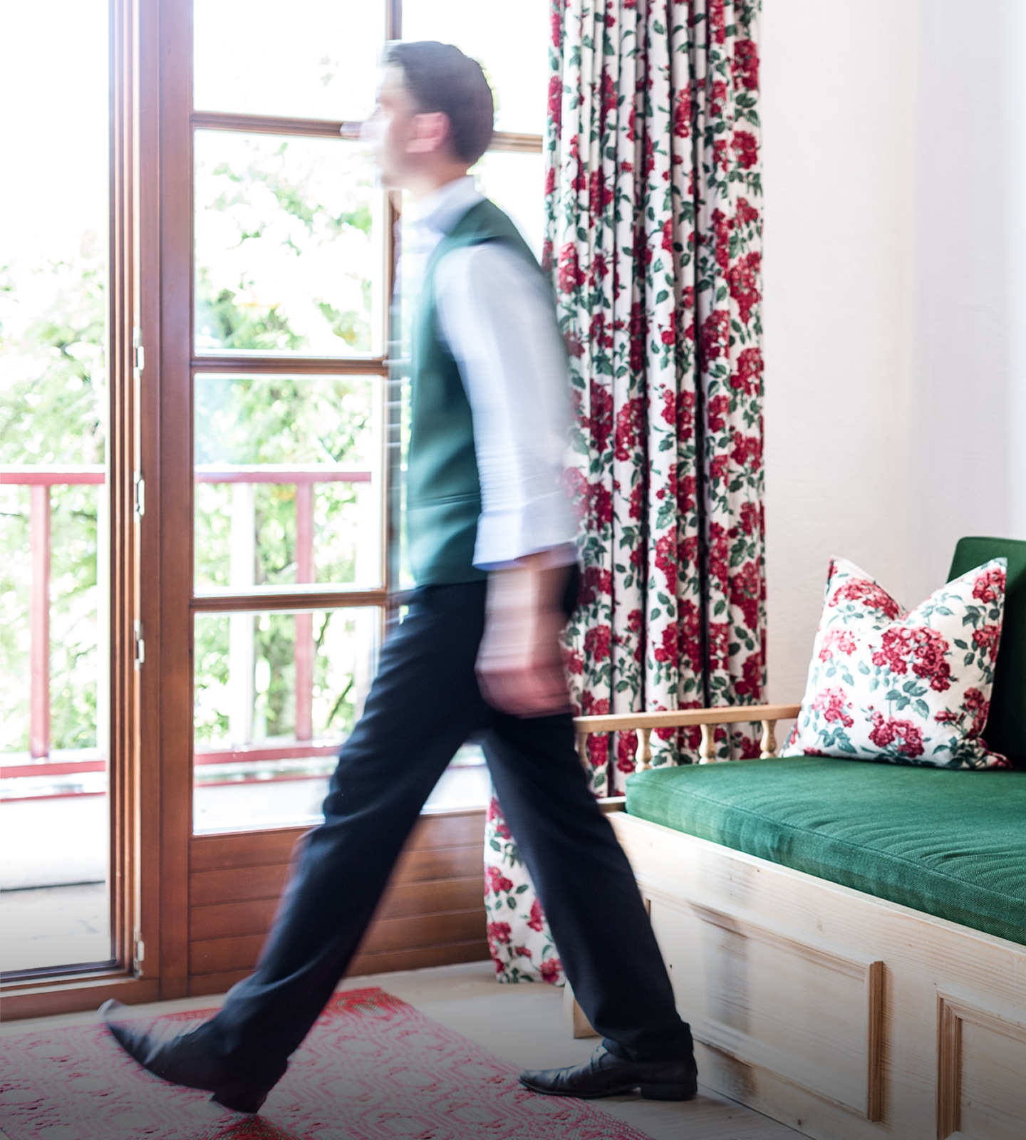 Ein Mann in Weste geht schwungvoll an einem Sofa mit Rosenkissen vorbei Richtung Balkon.