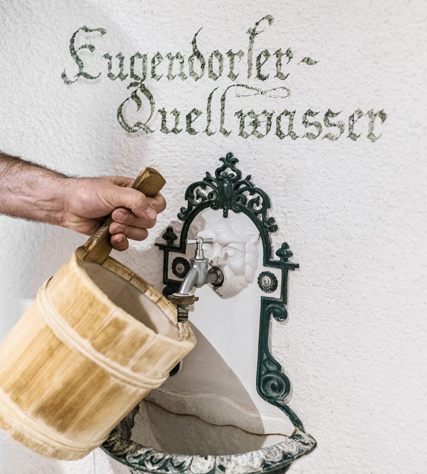 Ein Holzeimer wird unter einen Wandbrunnen mit der Aufschrift „Eugendorfer Quellwasser“ gehalten.