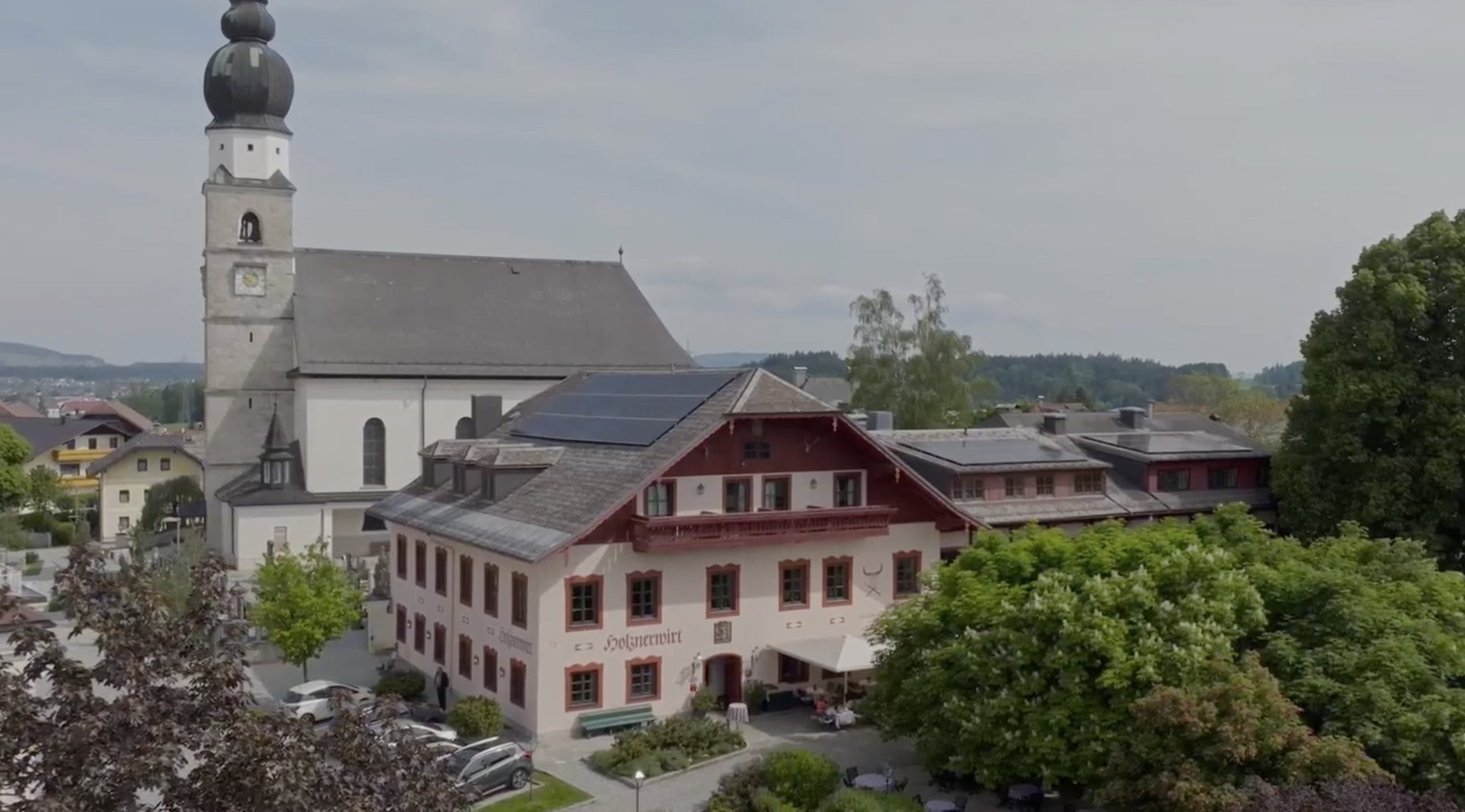 Der Gasthof Holznerwirt in Eugendorf mit Solaranlage auf dem Dach neben einer Kirche mit Zwiebelturm.