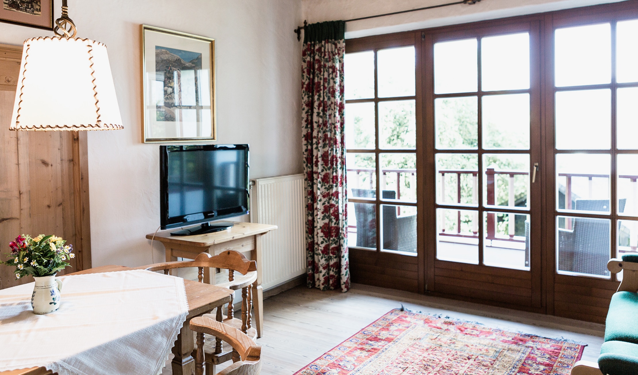 Rustikales Wohnzimmer mit Esstisch, Fernseher, großem Balkonfenster und gemustertem Teppich.