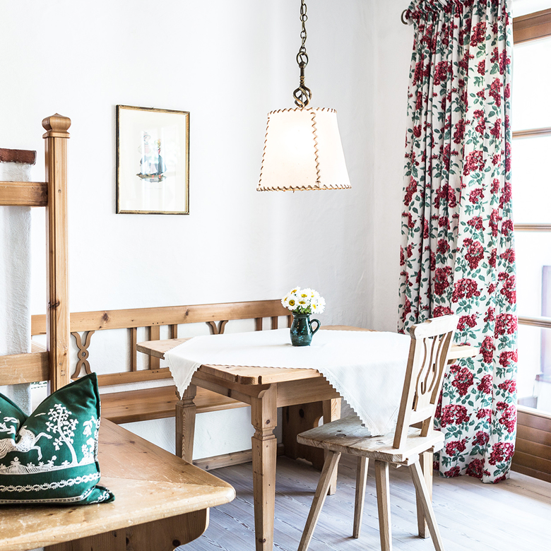 Gemütliche Essecke im Landhausstil mit Holztisch, Eckbank, Hängelampe und Vorhang mit rotem Blumenmuster.