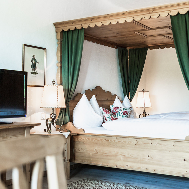 Rustikales Himmelbett aus Holz mit grünen Vorhängen und weißer Bettwäsche in einem gemütlichen Zimmer.