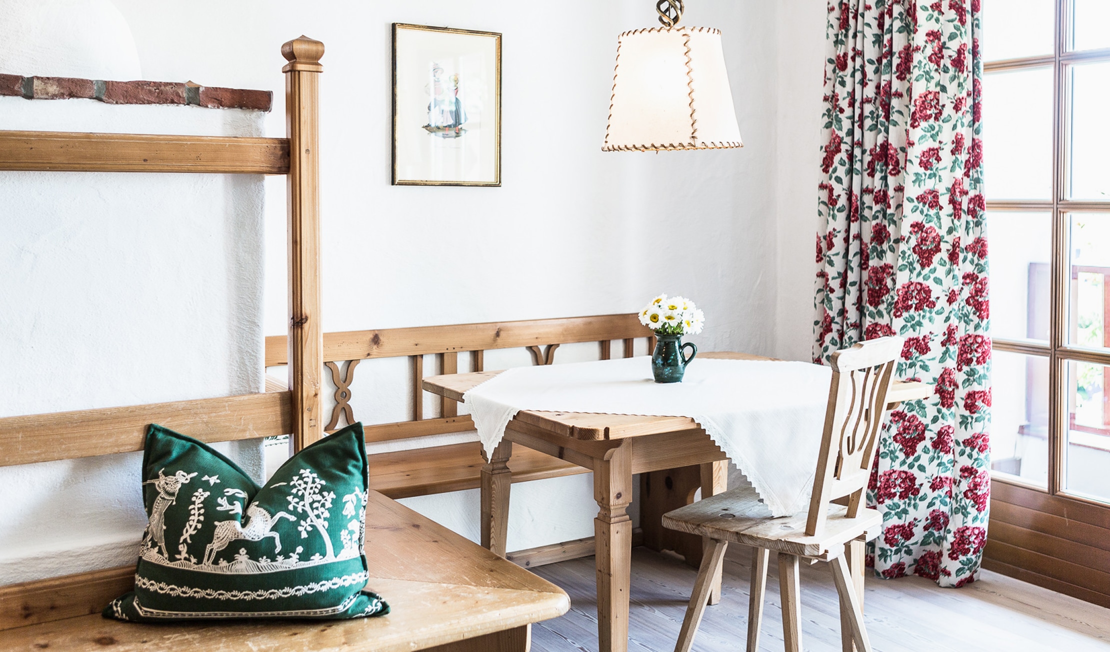 Gemütliche hölzerne Sitzecke mit Esstisch, Eckbank, besticktem Kissen und geblümtem Vorhang im Landhausstil.