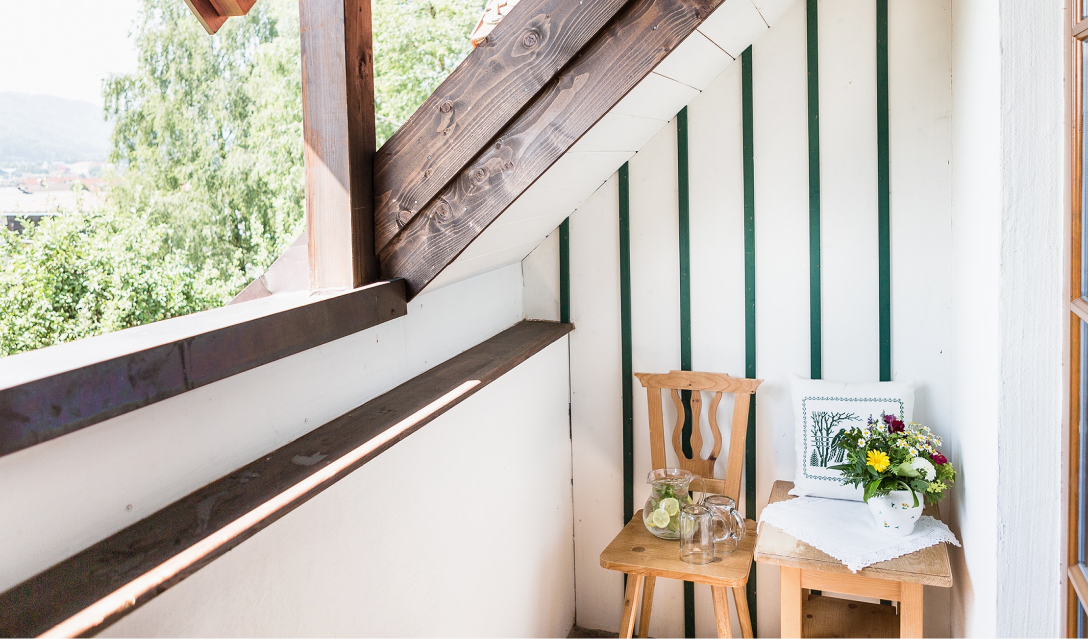 Gemütlicher Balkon mit Holzstühlen, Blumen, einer Karaffe Wasser und Blick ins Grüne unter einer Dachschräge.