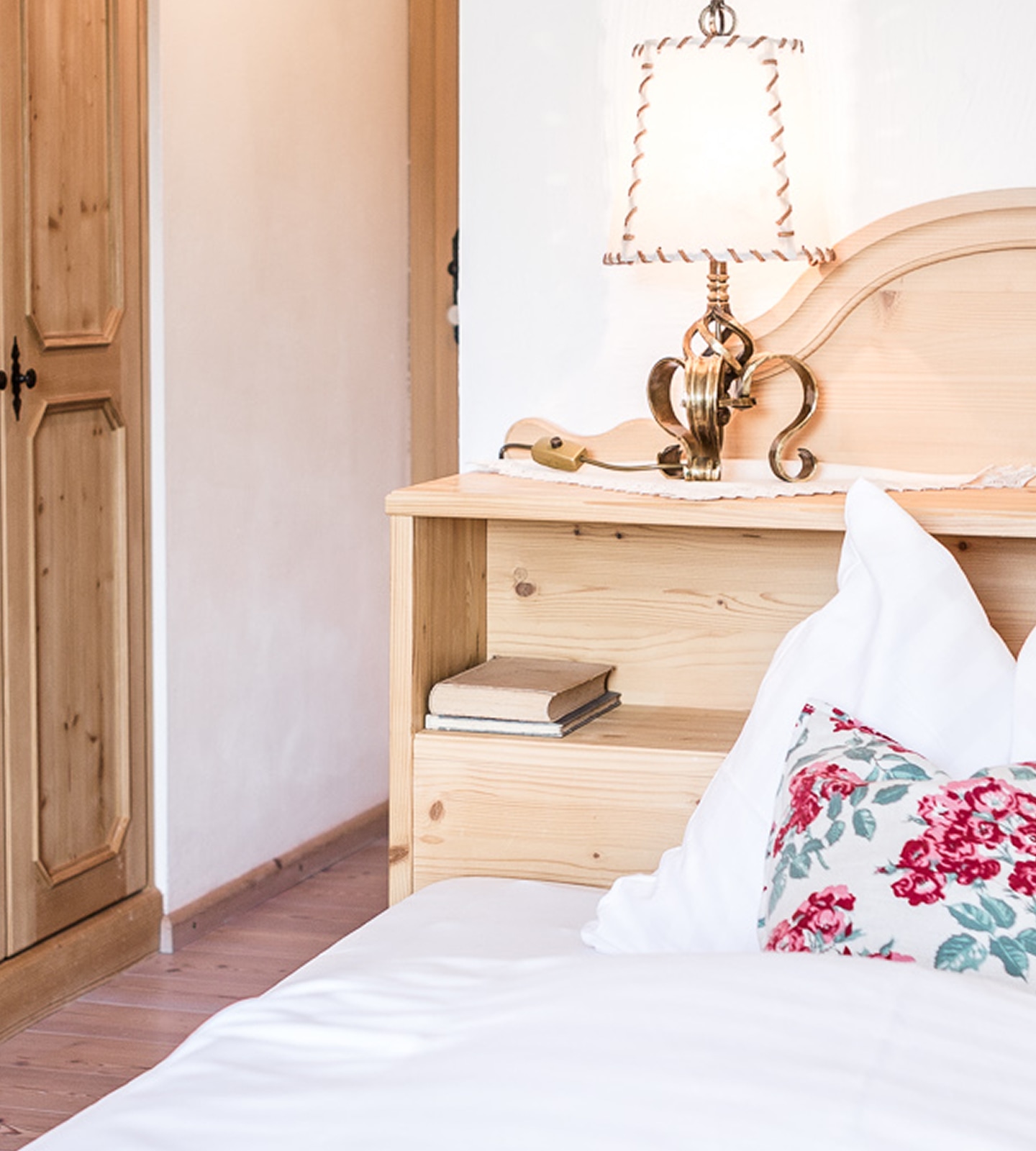 Rustikales Holzbett mit Nachttischlampe, Büchern im Fach und floral gemustertem Kissen im hellen Schlafzimmer.