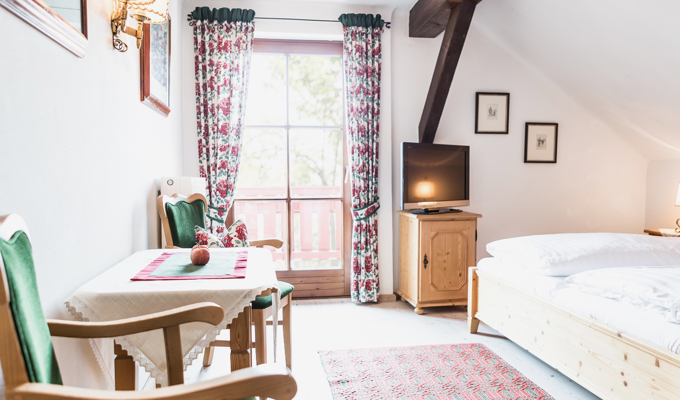Gemütliches Schlafzimmer im Landhausstil mit Holzbett, Sitzecke am Fenster, TV und floralen Vorhängen.