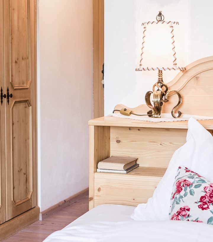 Schlafzimmer im Landhausstil mit hellem Holzbett, Nachttischlampe, weißer Bettwäsche und floralem Kissen.