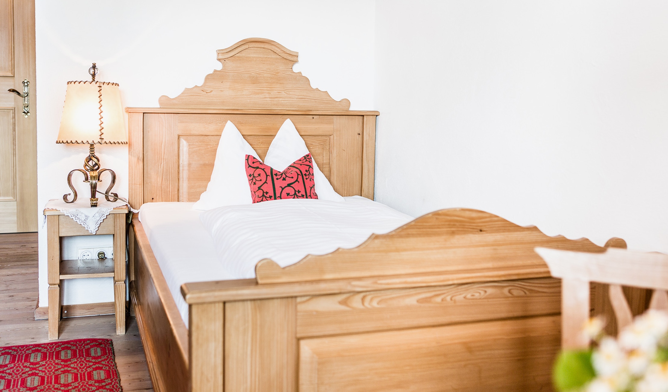 Rustikales Einzelbett aus hellem Holz mit weißer Bettwäsche, rotem Kissen und Nachttischlampe daneben.