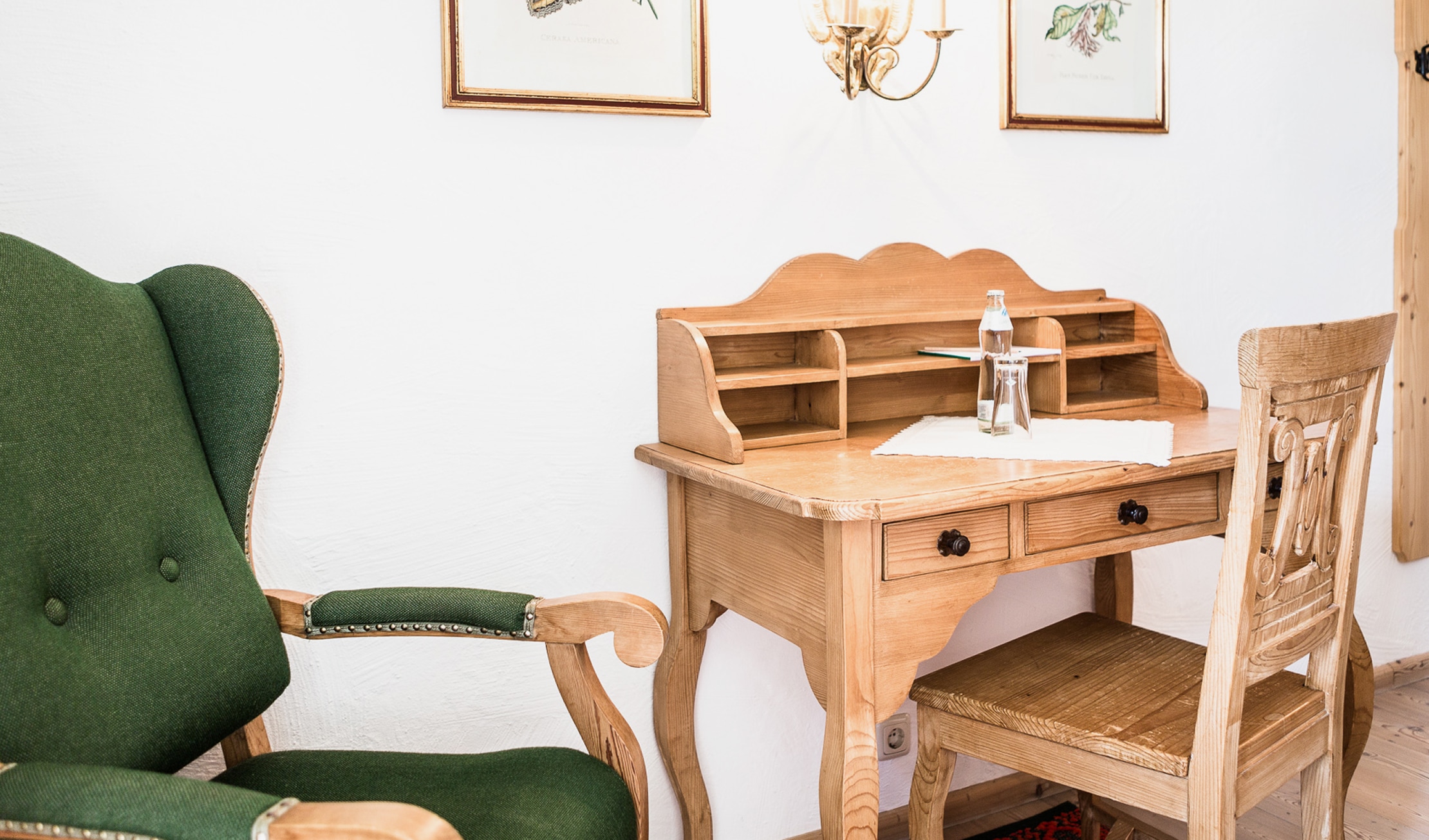 Klassischer Holzschreibtisch mit passendem Stuhl neben einem grünen Ohrensessel im Landhausstil.