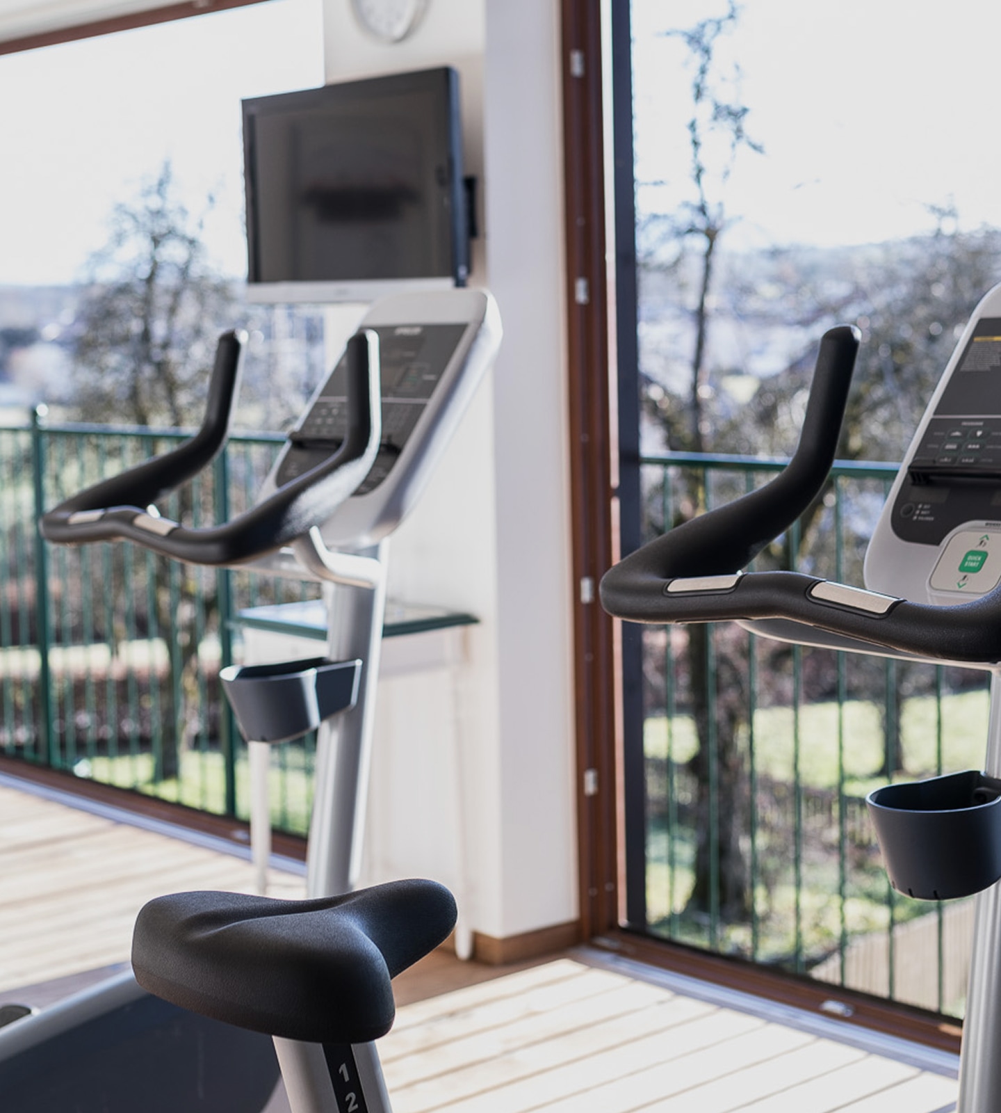 Zwei moderne Ergometer stehen in einem hellen Fitnessraum mit Blick ins Grüne durch große Fenster.
