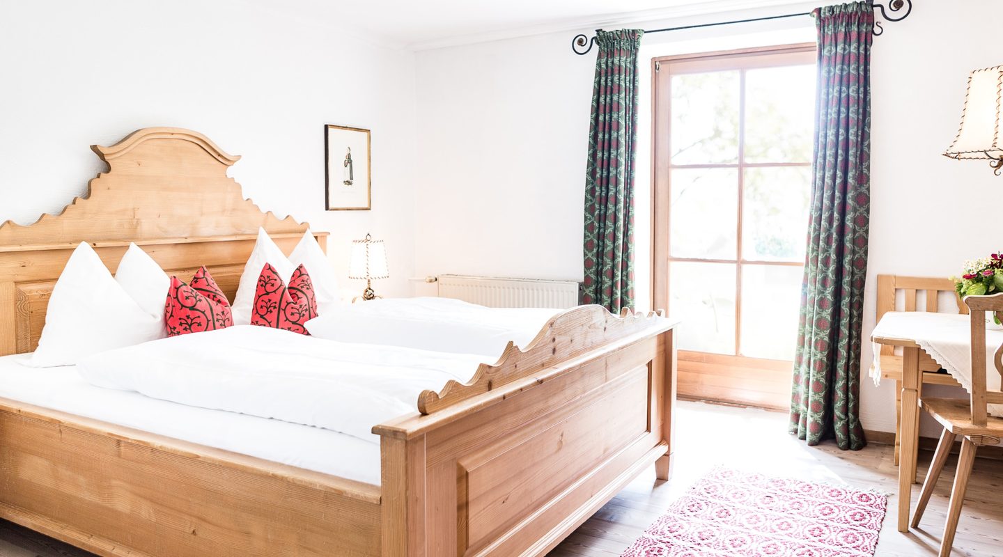 Gemütliches Schlafzimmer im Landhausstil mit Holzdoppelbett, roten Kissen, Balkontür und Holzmöbeln.