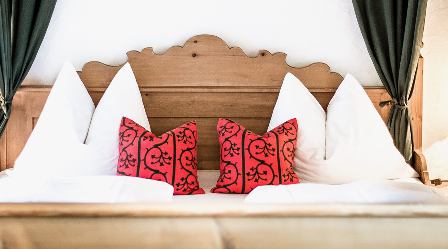 Holzbett mit weißen Kopfkissen und zwei verzierten roten Zierkissen im Landhausstil.
