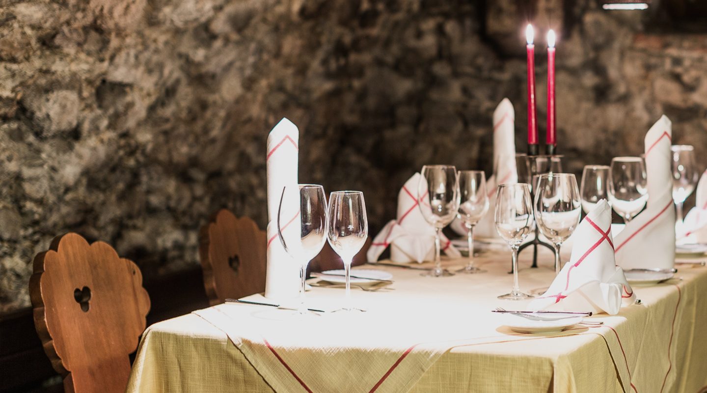 Festlich gedeckter Tisch in einem Kellergewölbe mit Weingläsern, Servietten und brennenden roten Kerzen.