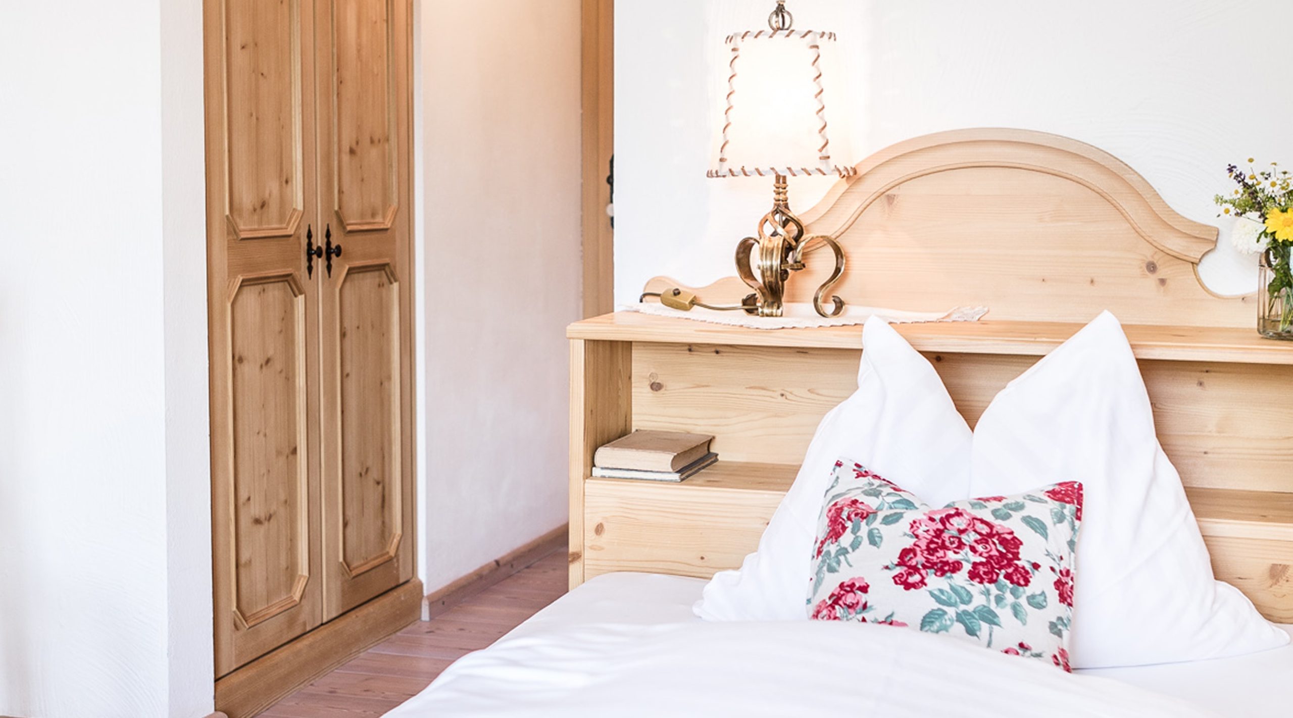 Gemütliches Schlafzimmer im Landhausstil mit Holzbett, weißem Kissen, floralem Dekokissen und Holzschrank.