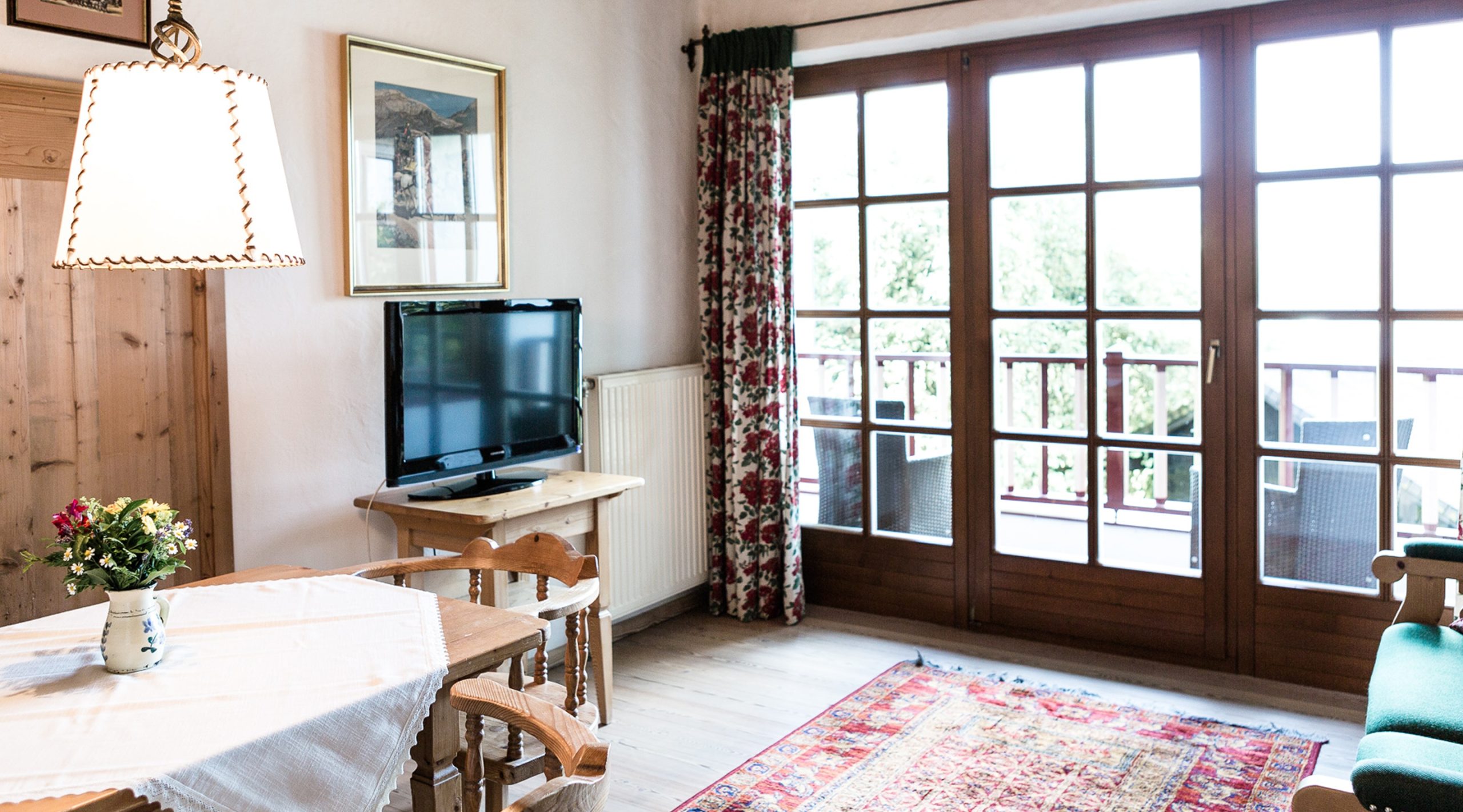 Gemütliches Wohnzimmer mit Holzmöbeln, Fernseher, großem Balkonfenster und buntem Teppich.