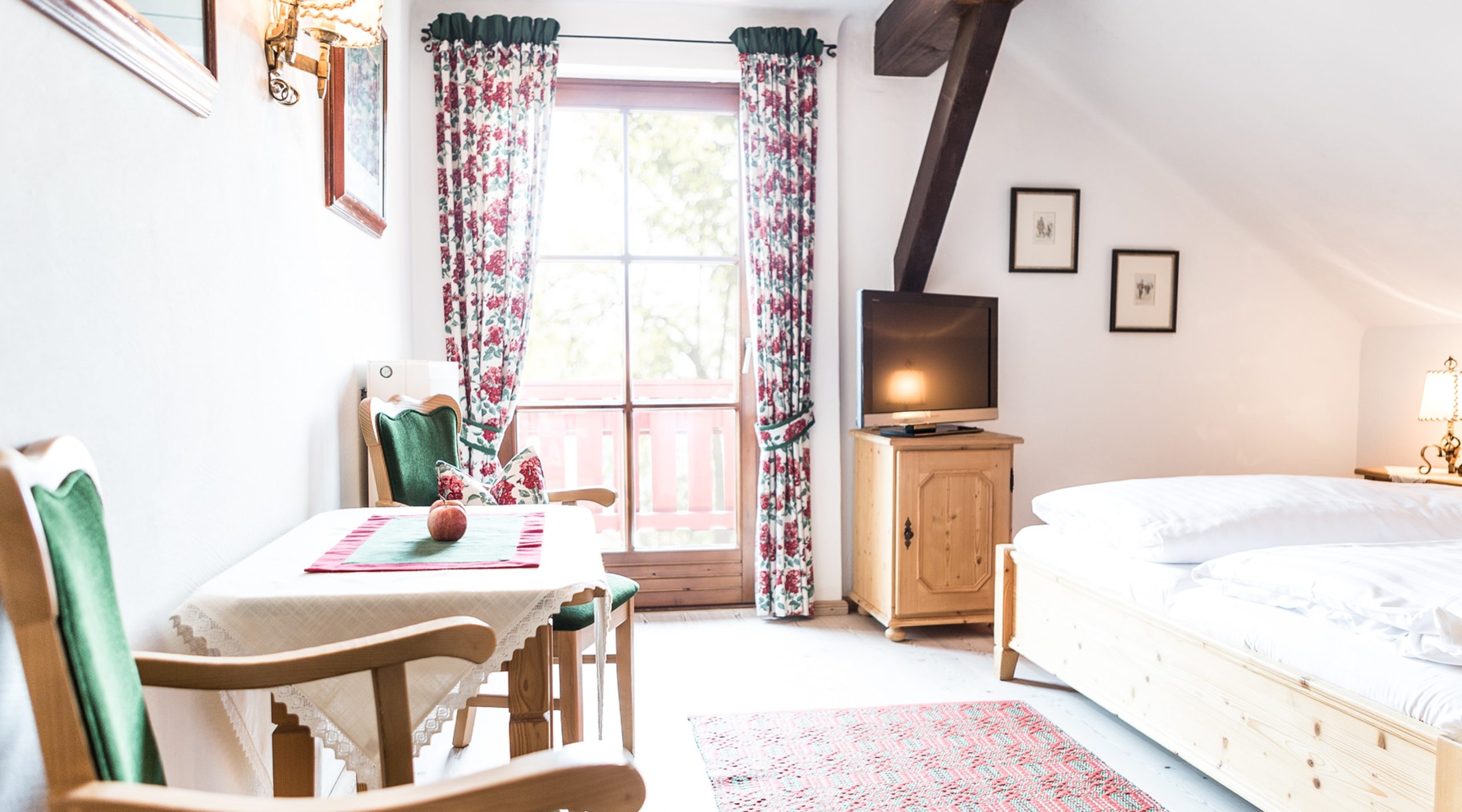 Gemütliches Schlafzimmer im Landhausstil mit Holzbett, Sitzecke, Fernseher und Balkontür mit geblümten Vorhängen.