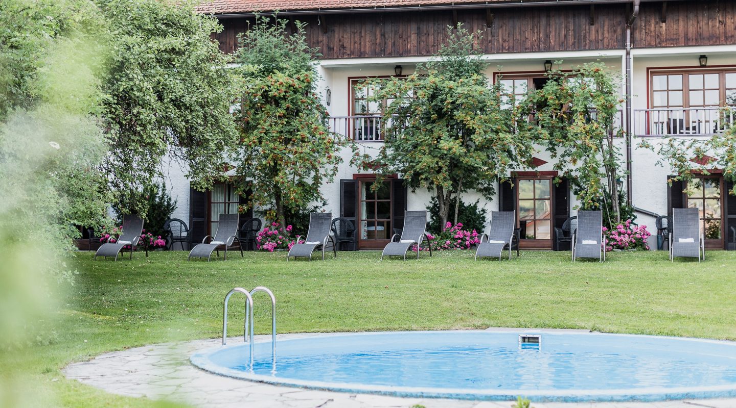 Ein runder Pool im Vordergrund einer grünen Wiese mit Liegestühlen vor einem Hotelgebäude im alpinen Stil.