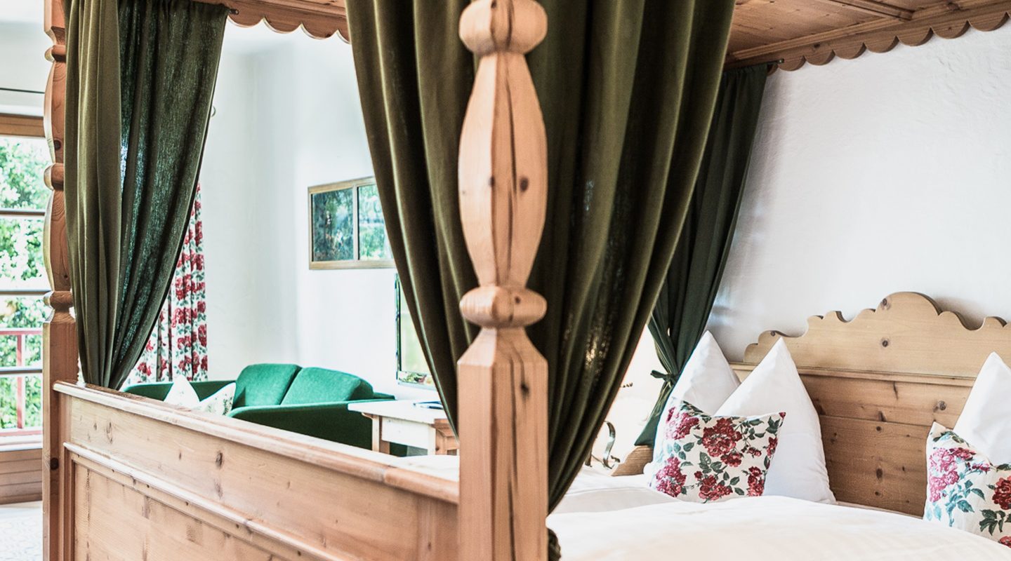 Rustikales Himmelbett aus Holz mit grünen Vorhängen und Blick auf ein grünes Sofa im Hintergrund.