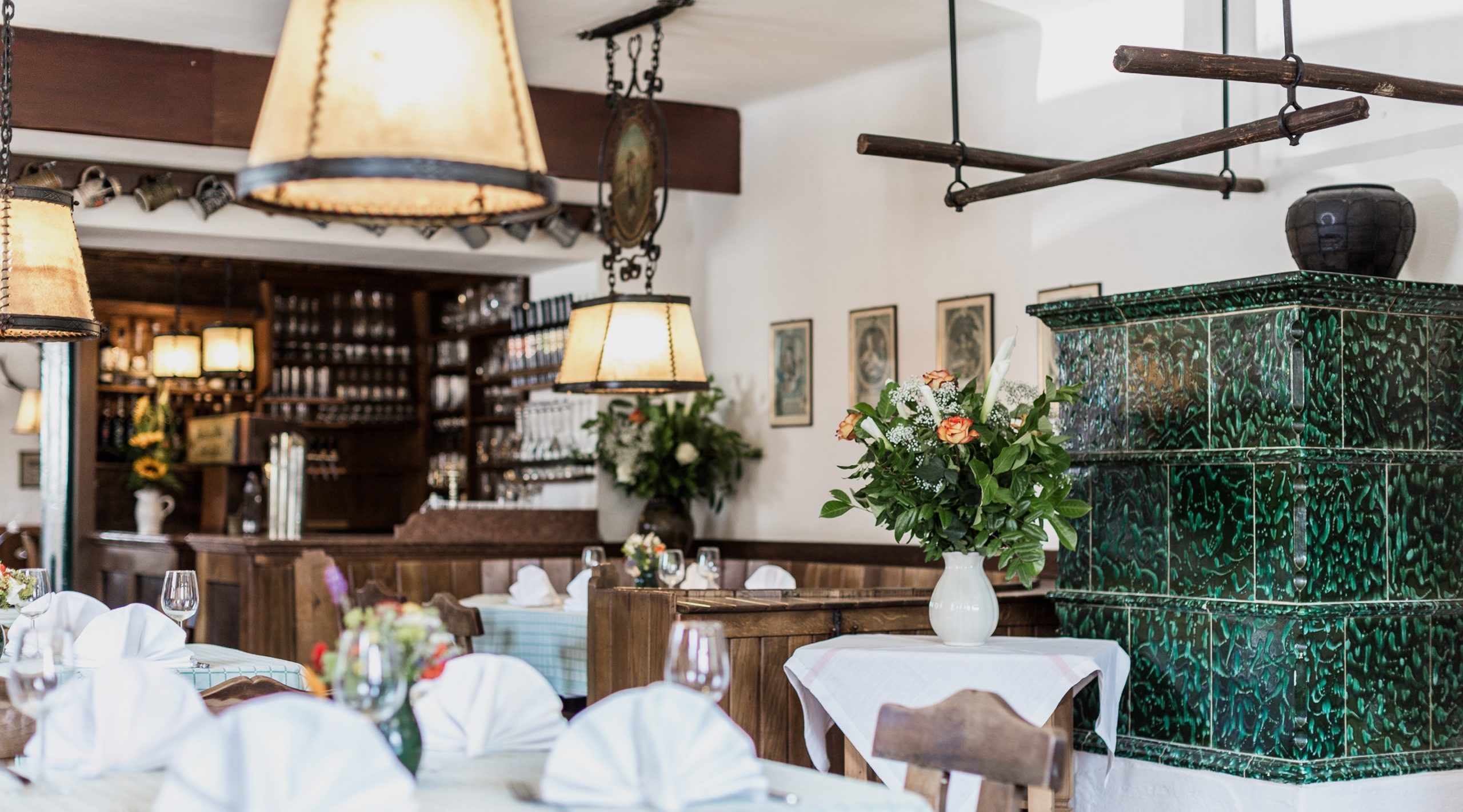 Einladender Restaurantbereich mit gedeckten Tischen, Blumen und einem traditionellen grünen Kachelofen.