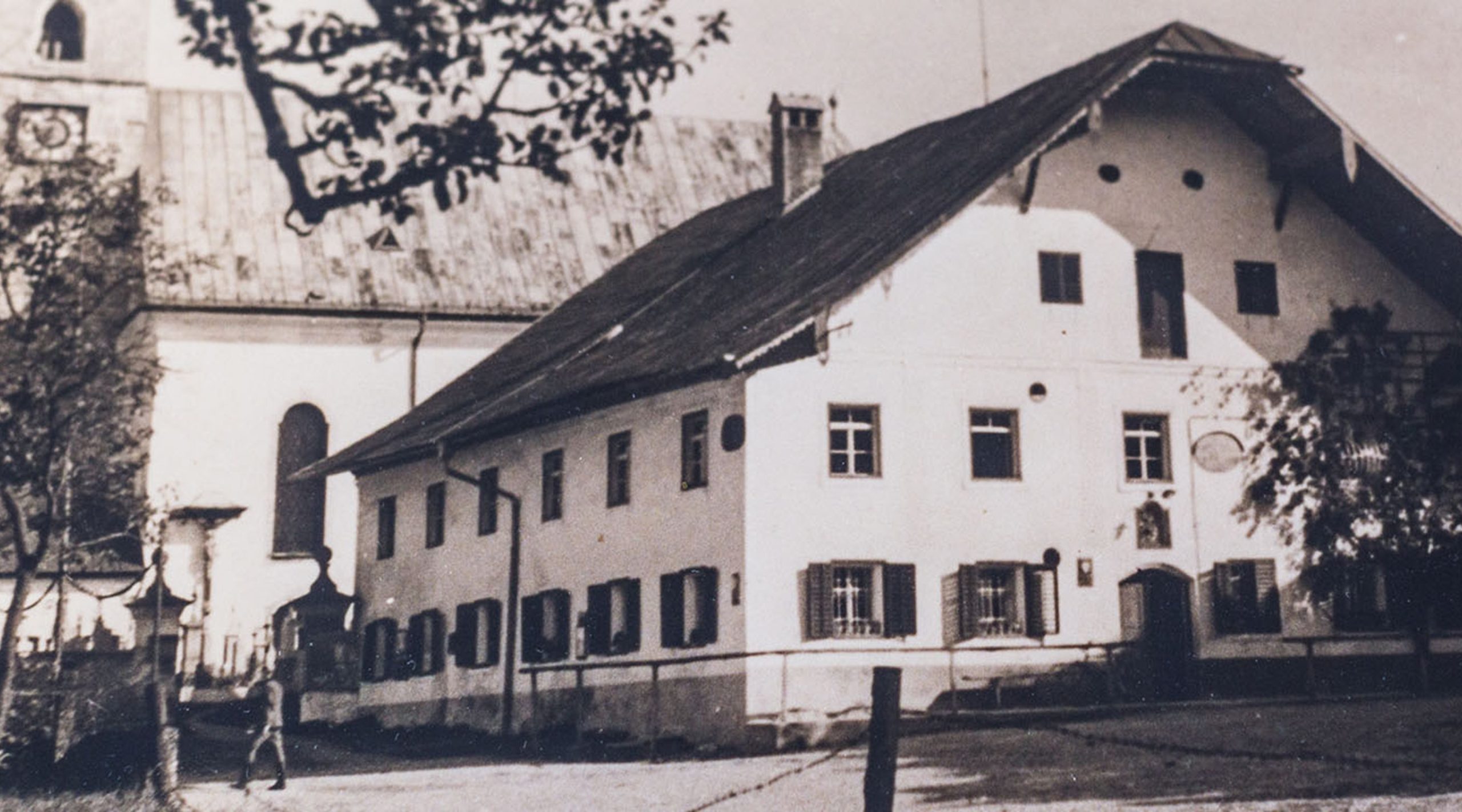 Historische Schwarz-Weiß-Aufnahme eines alten bayerischen Hauses neben einer Kirche mit Zifferblatt.