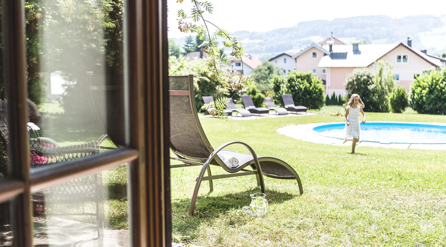 Blick aus dem Fenster auf einen Garten mit Liegestuhl, Pool und einem rennenden Mädchen.
