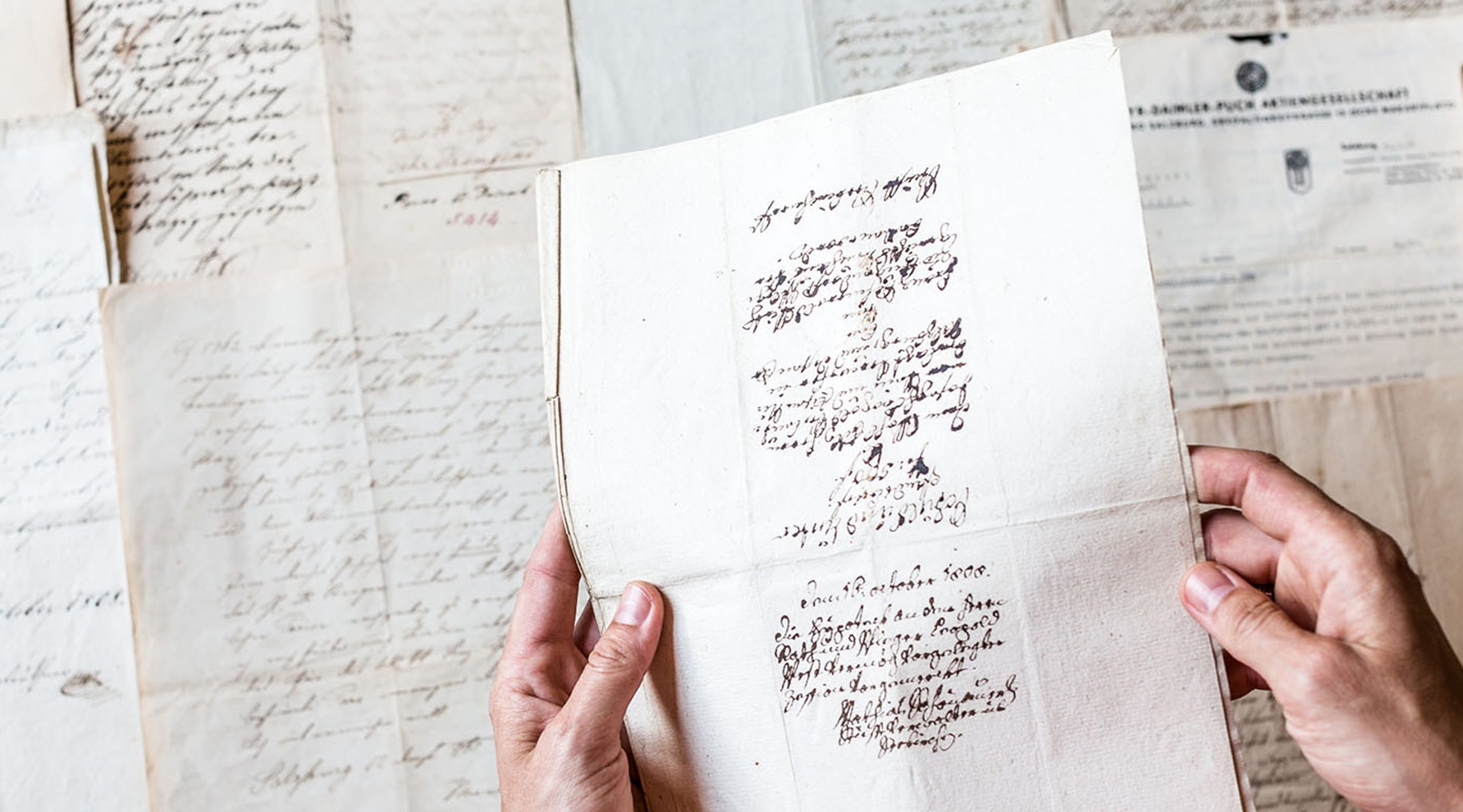 Hände halten ein altes, handschriftliches Dokument vor einem Hintergrund aus weiteren vergilbten Briefen.