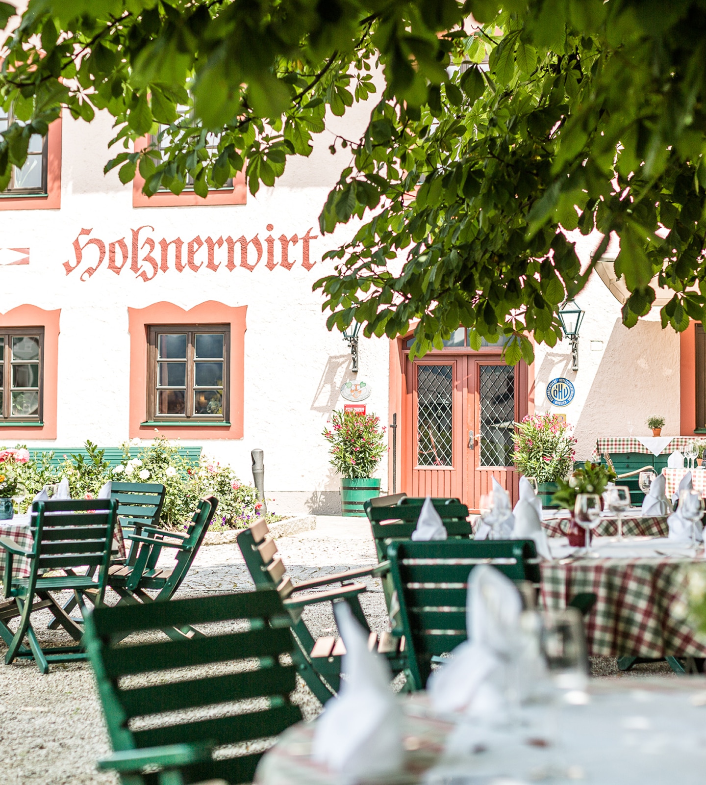 Der schattige Gastgarten des Hotels Holznerwirt mit gedeckten Tischen und grünen Holzstühlen vor der Hausfassade.