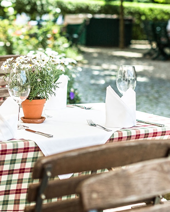 Gedeckter Tisch im Freien mit weißem Tischtuch, Blumen im Tontopf und Weingläsern in einem Gartenlokal.