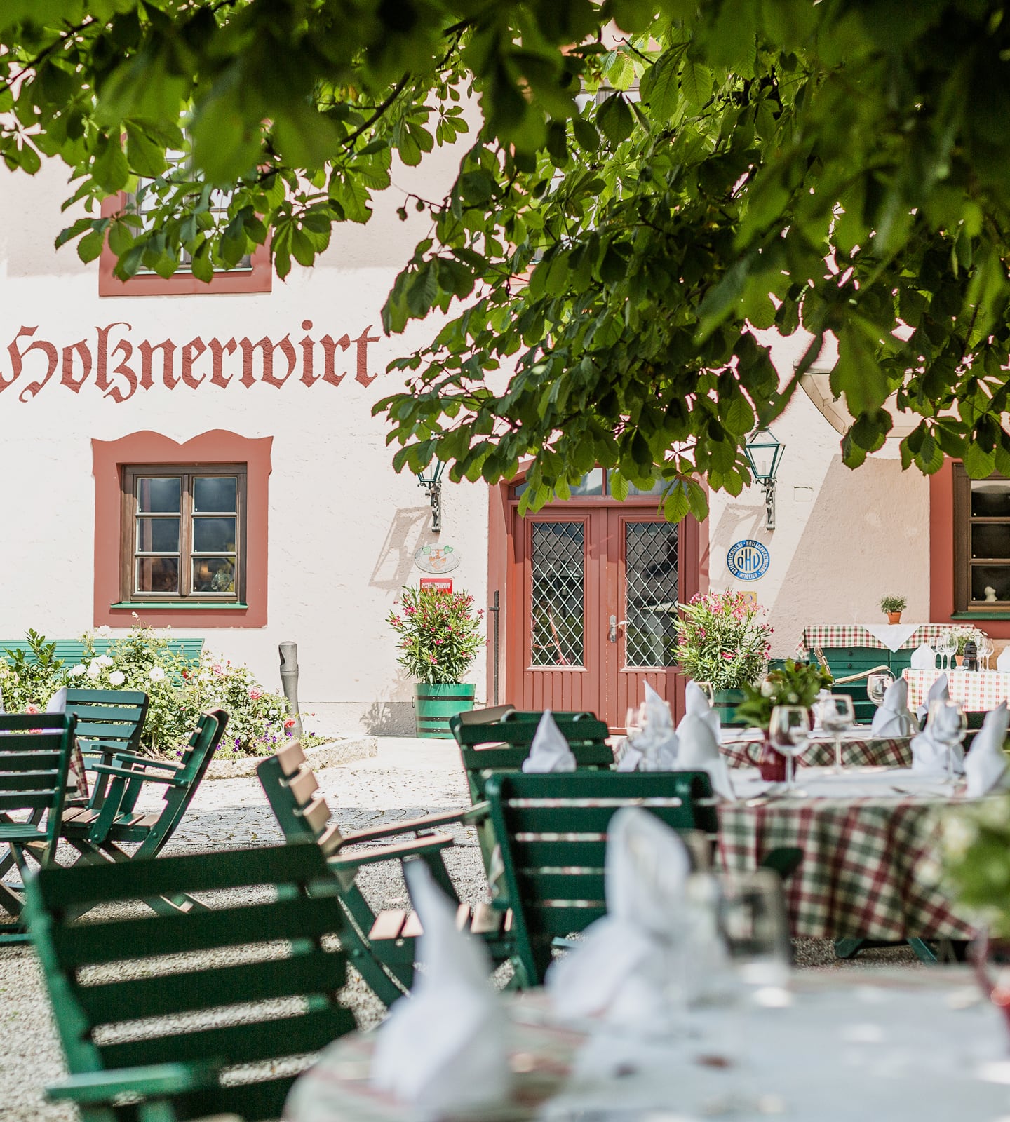 Biergarten des Holznerwirt mit gedeckten Tischen und grünen Holzstühlen unter schattigen Bäumen vor der Fassade.