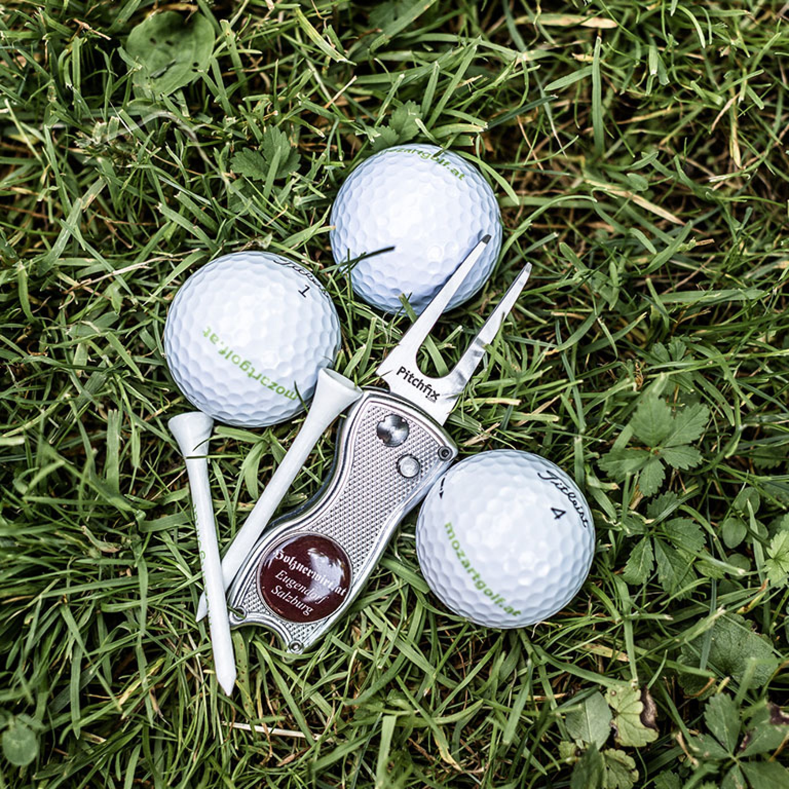 Drei Golfbälle mit mozartgolf.at-Aufdruck, zwei Tees und eine Pitchgabel im grünen Gras.
