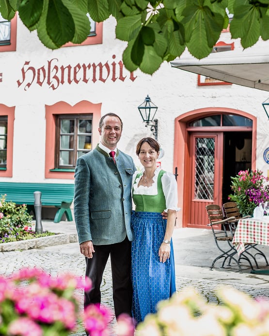 Ein lächelndes Paar in Tracht vor dem Gasthof „Holznerwirt“ im Freien.