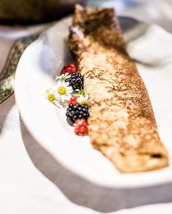 Ein goldbraun gebackener Crêpe auf einem weißen Teller, garniert mit Beeren und Kamillenblüten.