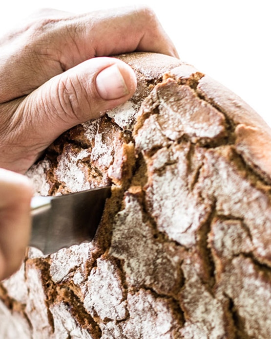Nahaufnahme von Händen, die ein frisches, rustikales Brot mit einem Messer anschneiden.