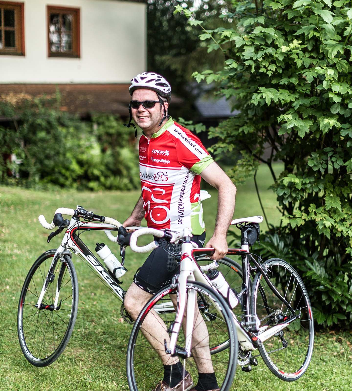 Ein lächelnder Rennradfahrer im rot-weißen Trikot hält zwei weiße Siga-Rennräder auf einer Wiese.