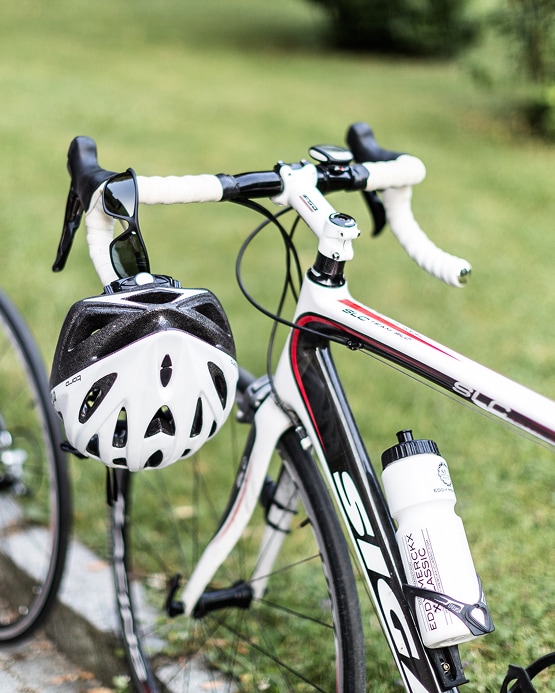 Rennrad mit weißem Helm am Lenker, einer Trinkflasche und Sonnenbrille vor grünem Hintergrund.
