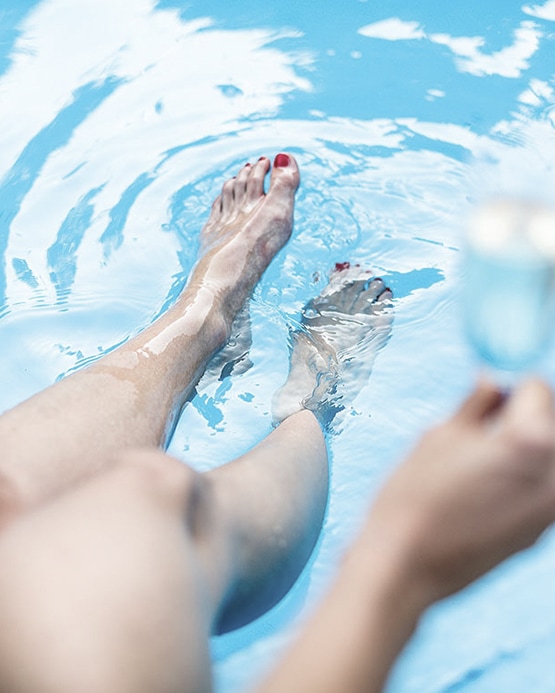 Beine mit rot lackierten Fußnägeln im blauen Poolwasser, im Vordergrund eine Hand mit Glas.