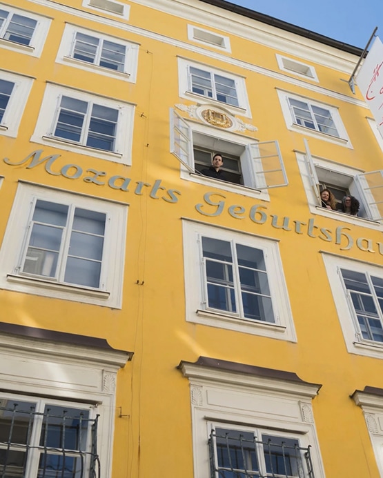 Die gelbe Fassade von Mozarts Geburtshaus in Salzburg mit Personen, die aus den Fenstern schauen.