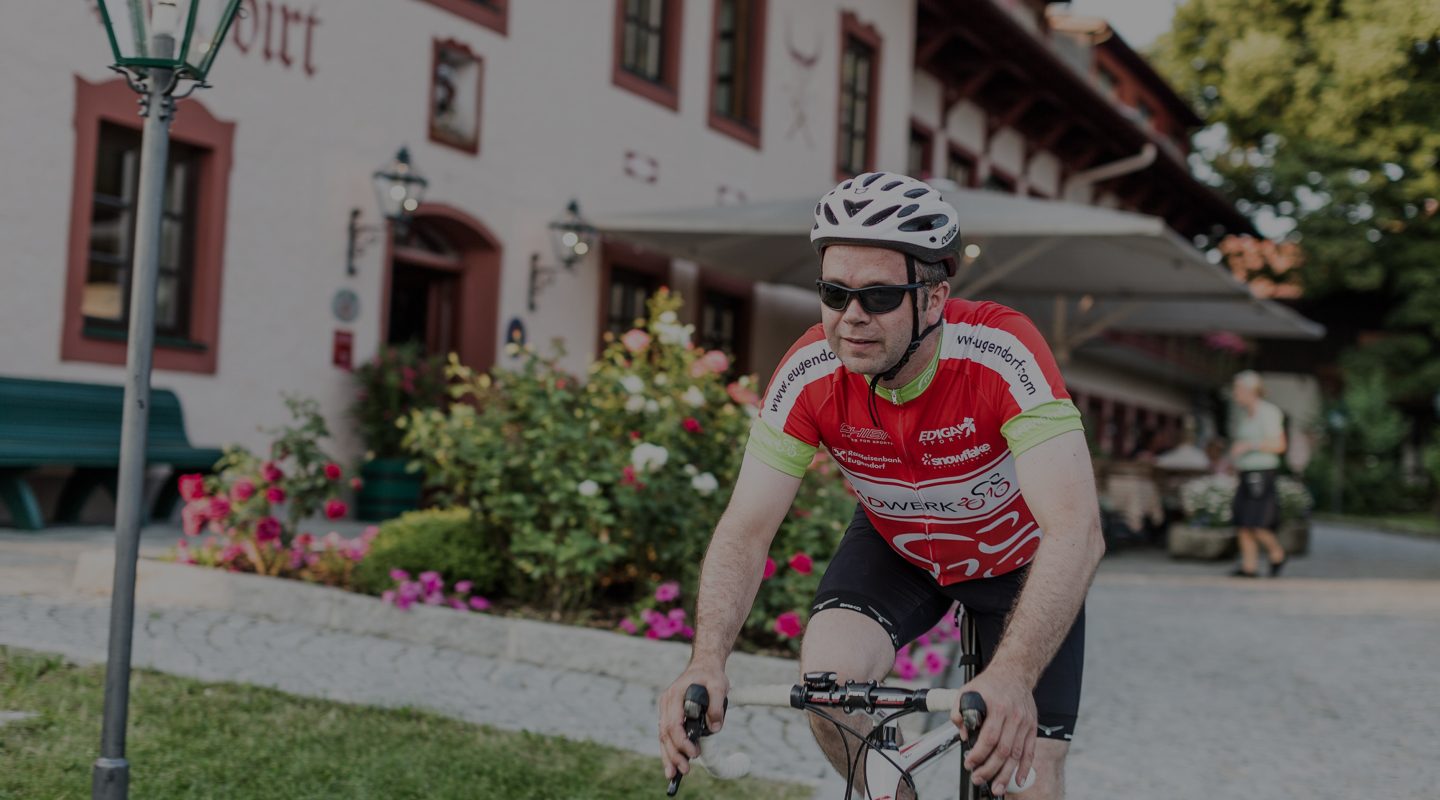 Ein Rennradfahrer mit Helm und Sonnenbrille fährt vor einem Hotelgebäude in Eugendorf.
