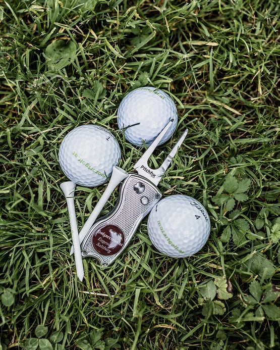 Drei Golfbälle, Tees und eine Pitchgabel mit der Aufschrift „Holznerwirt Eugendorf Salzburg“ im grünen Gras.